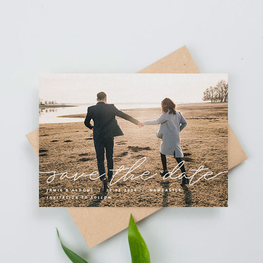 A landscape, modern wedding save the date card with photo. The save the date card has full-page photo with a handwritten “save the date” written across the bottom. The engaged couple’s names and the details of the wedding are printed below.