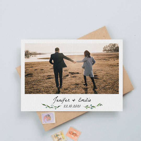 A simple, floral wedding save the date card with photos. It has small blue and green floral details either side of the printed date of a wedding. Above the date is the name of an engaged couple. At the top of the wedding save the date card is a photo of the engaged couple.