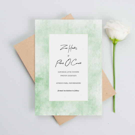 A rustic wedding save the date card with a worn green background. The centre of the save the date is white with black text detailing the particulars of an upcoming wedding.