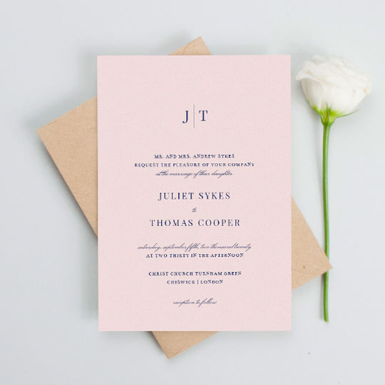 A classic pink wedding invitation with navy blue text. The top of the wedding invitation has a monogram of the bride and groom’s names.