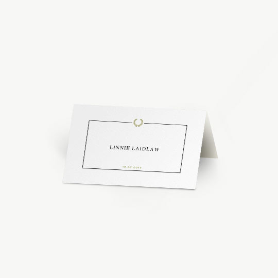 A white, tent place card, personalised with the details of a wedding guest. It has a simple black border with a gold, Greek wreath at the top-middle. The date of the wedding is printed at the bottom of the place card. It is a folded place card.