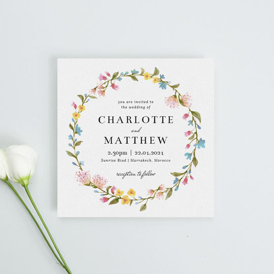 A floral wedding invitation made from a bright, summery floral wreath. The colour of the flowers on this wedding invitation are yellow, pink and blue, interspersed with green leaves.