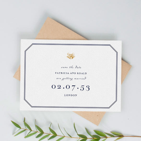 A vintage save the date wedding card. It has a classic two-lined border in navy blue, and gold wedding bells printed at the top of the card. The date of upcoming wedding is printed in a large, serif font. It is a classic save the date design.