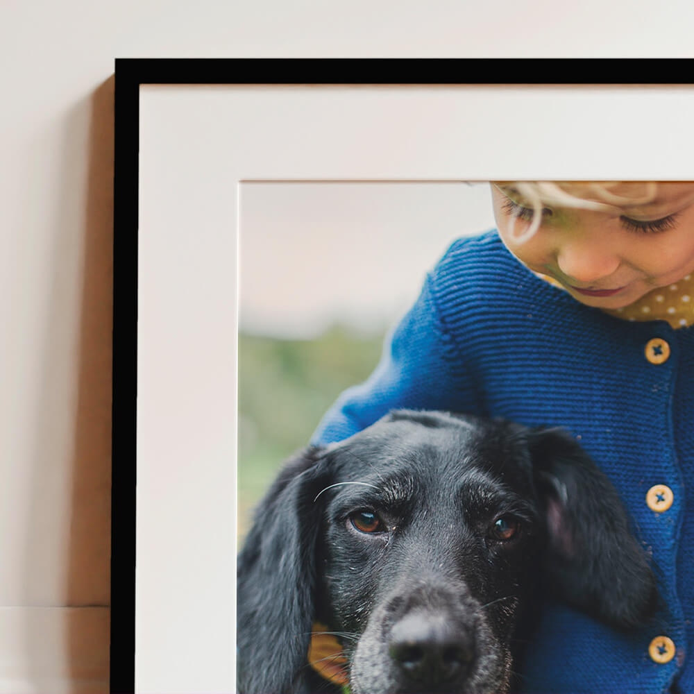 Framed and mounted photo print showing a young child in a blue jumper cuddling a black dog, displayed in a black frame with white mount