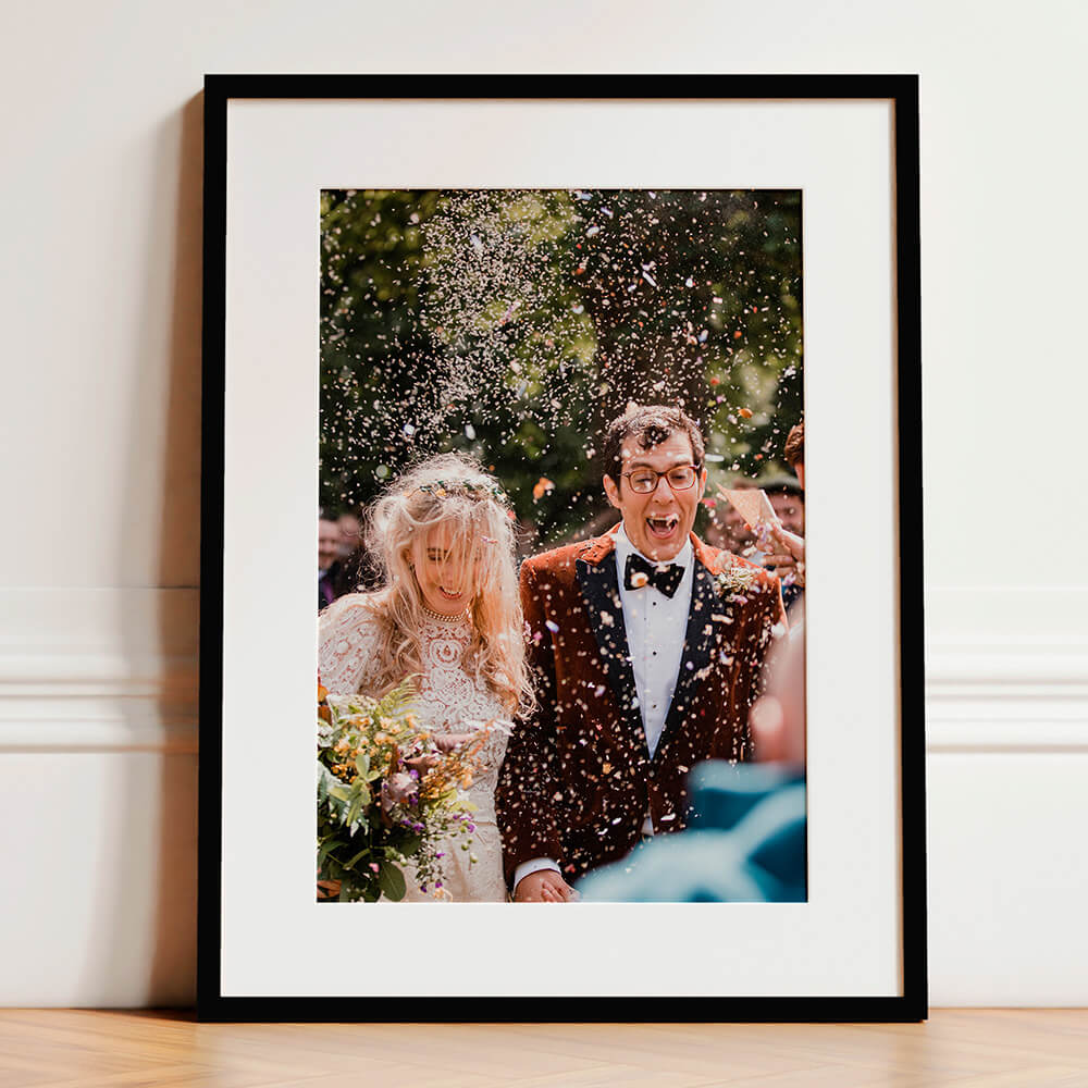 Framed and mounted wedding photo print showing a joyful bride and groom walking through confetti, displayed in a black frame with white mount